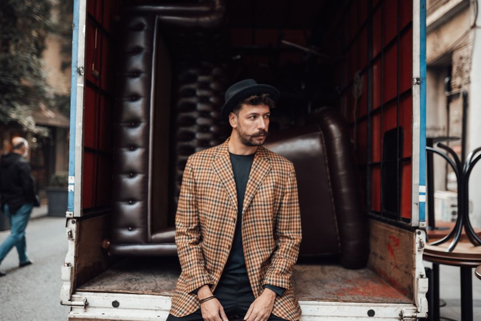 Fashionable bearded hipster male wearing black hat and jacket sitting in back of truck with furniture parked in city street