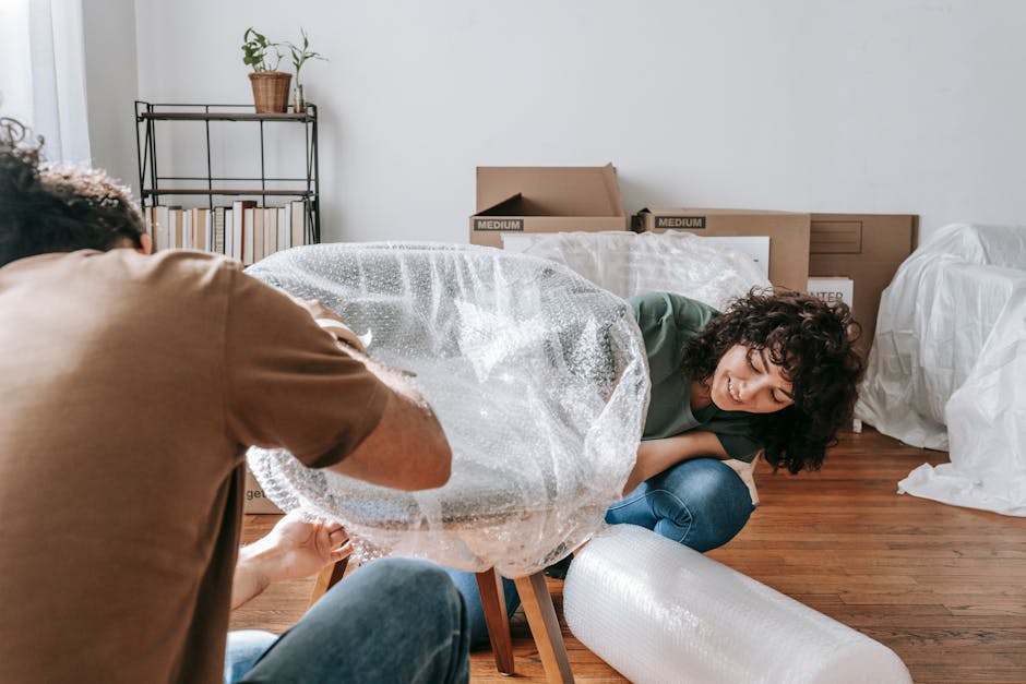 A couple carefully packing chairs with bubble wrap in a sunlit room during a move.